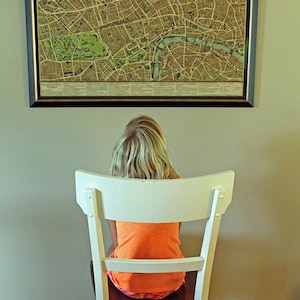 May include: A young person sits in a white chair facing a framed map of London. The map is in black and white with brown and green accents. The map is titled "The New Map of London".