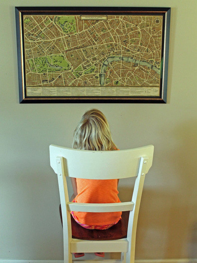 May include: A young person sits in a white chair facing a framed map of London. The map is in black and white with brown and green accents. The map is titled "The New Map of London".