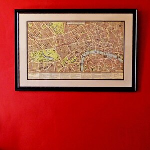 May include: A framed vintage map of London, England, with a detailed street map and a black frame. The map is hanging on a red wall.