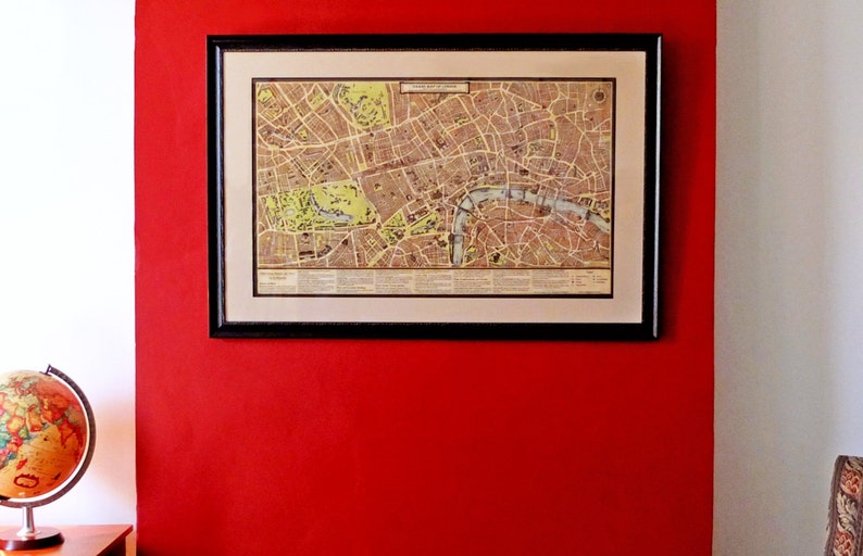 May include: A framed vintage map of London, England, with a detailed street map and a black frame. The map is hanging on a red wall.