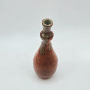 May include: A small, handcrafted ceramic vase with a bulbous body and a narrow neck. The vase features a reddish-brown glaze with darker brown and green accents. The vase is approximately 5 inches tall.