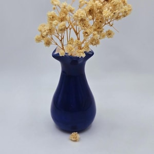 May include: A cobalt blue ceramic vase holds a bouquet of dried, beige flowers. The vase has a rounded body and a narrow neck. Several small flower heads are scattered around the base of the vase.