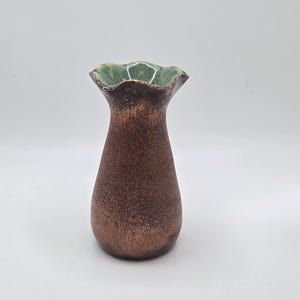 May include: A small, handcrafted vase with a textured, bronze-colored body and a ruffled, green-glazed rim. The vase has a unique, organic shape and is suitable for displaying small flowers or as a decorative accent.