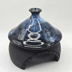 May include: A ceramic jar with a lid, featuring a dark blue and black glaze with white accents. The jar sits on a black base with an arched design. The lid has a small, rounded handle.