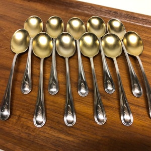 May include: A set of twelve silver spoons with gold-plated bowls. The spoons are arranged in a row on a wooden surface.