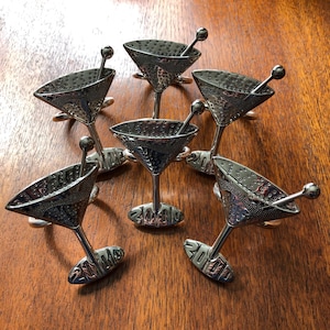 Peut inclure: Six ronds de serviette en forme de verre à martini argentés avec des agitateurs. Chaque anneau a un bol texturé et une base avec le numéro "2000". Les verres à martini sont disposés sur une surface en bois.