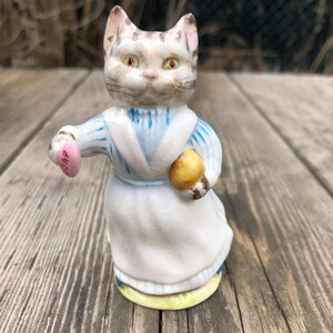 May include: A ceramic cat figurine in a blue and white striped dress and white apron. The cat holds a pink soap in one paw and a yellow object in the other. The figurine has a yellow base. Set against a wooden background.