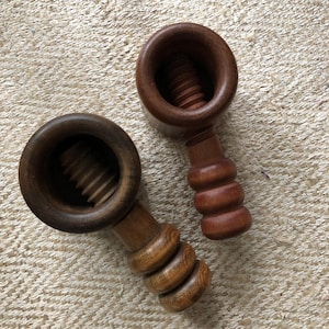 Può includere: Due schiaccianoci in legno con meccanismo a vite. Gli schiaccianoci sono realizzati in legno marrone scuro e chiaro. I manici hanno un design arrotondato. Gli schiaccianoci sono su una superficie beige strutturata.