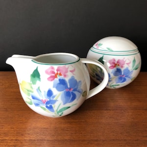 Peut inclure: Un ensemble de crémier et de sucrier en porcelaine blanche avec un motif floral. Le crémier a un corps arrondi et une anse. Le sucrier est rond et possède un couvercle. Les deux pièces présentent une bordure verte et un motif floral avec des fleurs roses, bleues et jaunes.