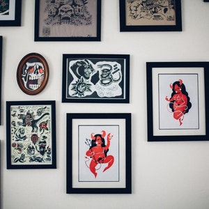 May include: A white wall with a collection of framed artwork. The frames are black and various sizes. The artwork includes a group photo of people, a skull, a snake, a devil, and a cartoon illustration of a devil.