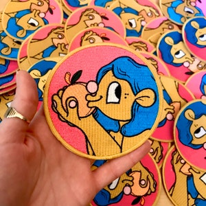 May include: A round embroidered patch with a pink background and a yellow outline. The patch features a cartoon character with blue hair, a peach-coloured face, and a large eye. The character is holding a peach in their hand and has a pink blush on their cheek.