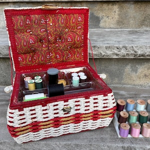 May include: A rectangular woven sewing basket with a red and white pattern, featuring a hinged lid with a red paisley fabric interior and a clear plastic tray insert containing various spools of thread and a separate white plastic thread rack.