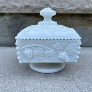 May include: A white, vintage-style candy dish with a lid and pedestal base. The dish features embossed grape and leaf designs on the sides and lid. The lid has a handle. The dish is made of opaque white glass.