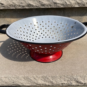 May include: A red and white enamel colander with black handles. The colander has a red base and a white interior with numerous small holes for draining. The handles are black and curved. This kitchenware item is designed for straining food.