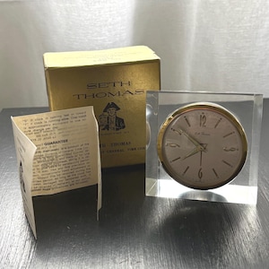 May include: A Seth Thomas analog clock encased in a clear, rectangular acrylic block. The clock face is white with gold-tone numerals and hands. Included is a gold-colored cardboard box with the brand name and a printed product guarantee pamphlet.