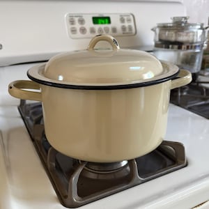 May include: A cream-colored enamel stockpot with a matching lid and two side handles. The pot features a smooth, glossy finish with a thin black rim around the top edge of the base and the lid. The lid has a small, arched handle on top.