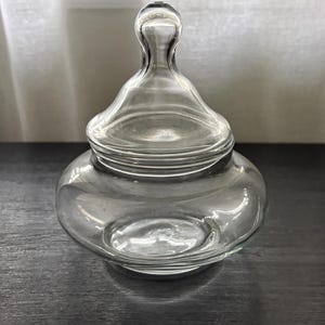 May include: A clear glass candy dish with a rounded base and a matching removable lid. The lid features a smooth, bulbous finial handle at the top. The container has a wide, curved body and a circular opening, crafted from transparent glass.