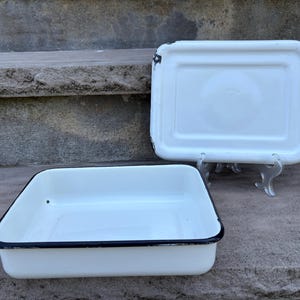 May include: A white rectangular enamel baking pan with a black rim and a matching white enamel lid featuring a black rim. The lid is displayed upright on a small clear stand behind the open pan, showing a smooth surface and worn edges.