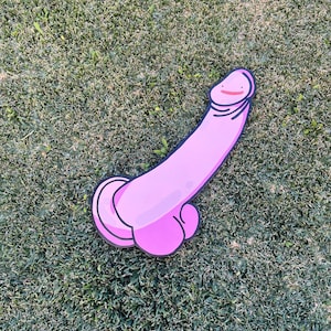 Pode incluir: Um grande recorte de falo rosa em estilo cartoon com um rosto sorridente na cabeça. O recorte tem um contorno preto e está sobre um leito de relva verde.