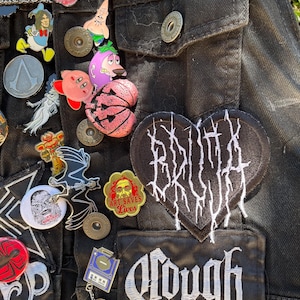 May include: A black denim jacket with a variety of patches and pins. The patches include a heart-shaped patch with the text "BRUTAL TRUTH" in white, a patch with the text "OUGH" in white, and a patch with the text "ART SAVES LIVES" in red and white. The pins include a variety of cartoon characters, a skull, and a bat.