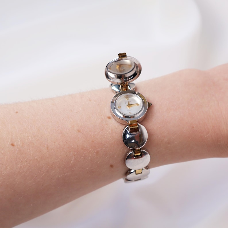Puede incluir: Un reloj plateado y dorado con un dise&ntilde;o &uacute;nico. El reloj presenta una serie de eslabones circulares, con tres esferas visibles. Una esfera est&aacute; abierta, revelando las manecillas. El reloj se lleva en la mu&ntilde;eca.
