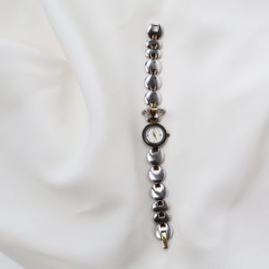 Puede incluir: Un reloj de pulsera plateado con una esfera redonda negra y una esfera blanca. La correa del reloj est&aacute; compuesta por eslabones circulares conectados. El reloj tiene detalles dorados y est&aacute; sobre un fondo blanco.