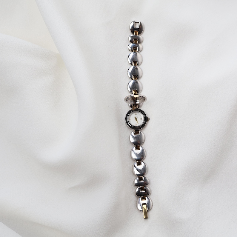Puede incluir: Un reloj de pulsera plateado con una esfera redonda negra y una esfera blanca. La correa del reloj est&aacute; compuesta por eslabones circulares conectados. El reloj tiene detalles dorados y est&aacute; sobre un fondo blanco.