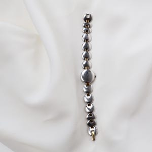 Puede incluir: Un reloj de pulsera plateado con esfera redonda y correa de eslabones. La correa presenta una serie de eslabones circulares y rectangulares. El reloj tiene detalles dorados en el cierre y las conexiones de la correa. Sobre fondo blanco.