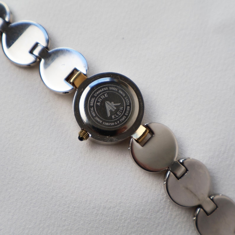 Puede incluir: Reloj Anne Klein con una correa de metal plateado y una esfera redonda. La esfera es negra con detalles dorados. La correa presenta una serie de eslabones circulares conectados. La parte trasera del reloj es de acero inoxidable.
