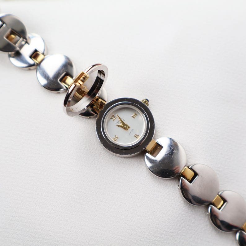 Puede incluir: Un reloj plateado y dorado con esfera blanca y n&uacute;meros romanos. El reloj tiene una esfera circular y est&aacute; unido a una pulsera con eslabones circulares. El reloj est&aacute; abierto, revelando la esfera.