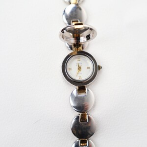 Puede incluir: Reloj Anne Klein plateado con esfera blanca y detalles dorados. El reloj presenta un dise&ntilde;o &uacute;nico de medall&oacute;n que se abre. La correa est&aacute; compuesta por eslabones circulares plateados y dorados conectados.