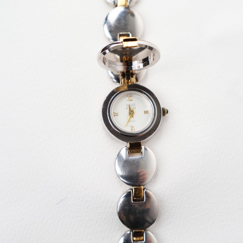 Puede incluir: Reloj Anne Klein plateado con esfera blanca y detalles dorados. El reloj presenta un dise&ntilde;o &uacute;nico de medall&oacute;n que se abre. La correa est&aacute; compuesta por eslabones circulares plateados y dorados conectados.