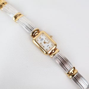 May include: A gold and silver Anne Klein wristwatch with a rectangular face and a white dial. The watch has gold accents and a silver band. The watch face displays the Anne Klein logo and the word "Swiss".