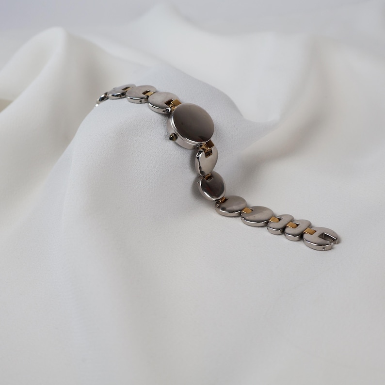 Puede incluir: Un reloj de pulsera plateado con una esfera circular y una correa de eslabones. La correa presenta una serie de eslabones planos y redondos con detalles dorados. El reloj se muestra sobre una superficie blanca texturizada.