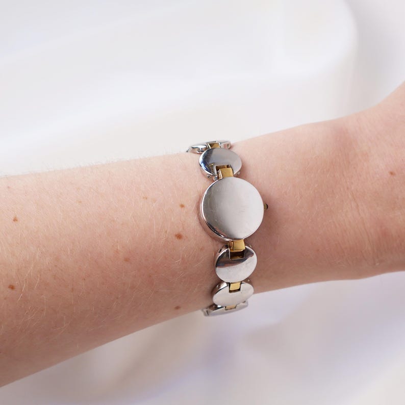 Puede incluir: Un reloj de pulsera plateado y dorado con esfera circular y correa de eslabones. El reloj presenta una esfera plateada con detalles dorados en los eslabones de la correa. El reloj se lleva en la mu&ntilde;eca.