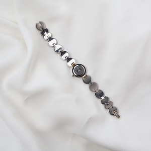 Puede incluir: Reloj de pulsera plateado con esfera circular y una correa compuesta por eslabones redondos y planos conectados. La esfera del reloj tiene un fondo oscuro con un logotipo. El reloj se muestra sobre una tela blanca texturizada.