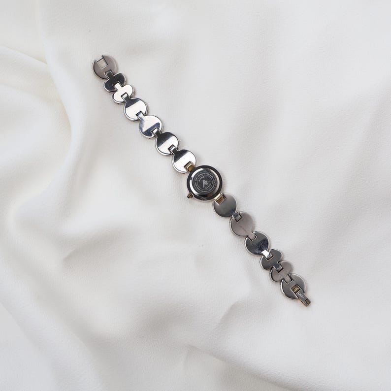 Puede incluir: Reloj de pulsera plateado con esfera circular y una correa compuesta por eslabones redondos y planos conectados. La esfera del reloj tiene un fondo oscuro con un logotipo. El reloj se muestra sobre una tela blanca texturizada.