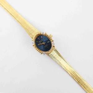 May include: A gold-tone wristwatch featuring an oval-shaped deep blue dial with gold hands and markers. The watch case is surrounded by a twisted gold-tone border with small clear stones. It has a textured, flexible gold-tone metal link strap.