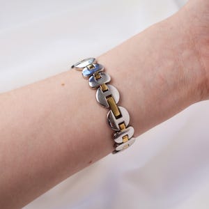 Puede incluir: Una pulsera plateada y dorada con un dise&ntilde;o de eslabones. La pulsera presenta eslabones circulares plateados conectados por barras rectangulares doradas. La pulsera se lleva en la mu&ntilde;eca.