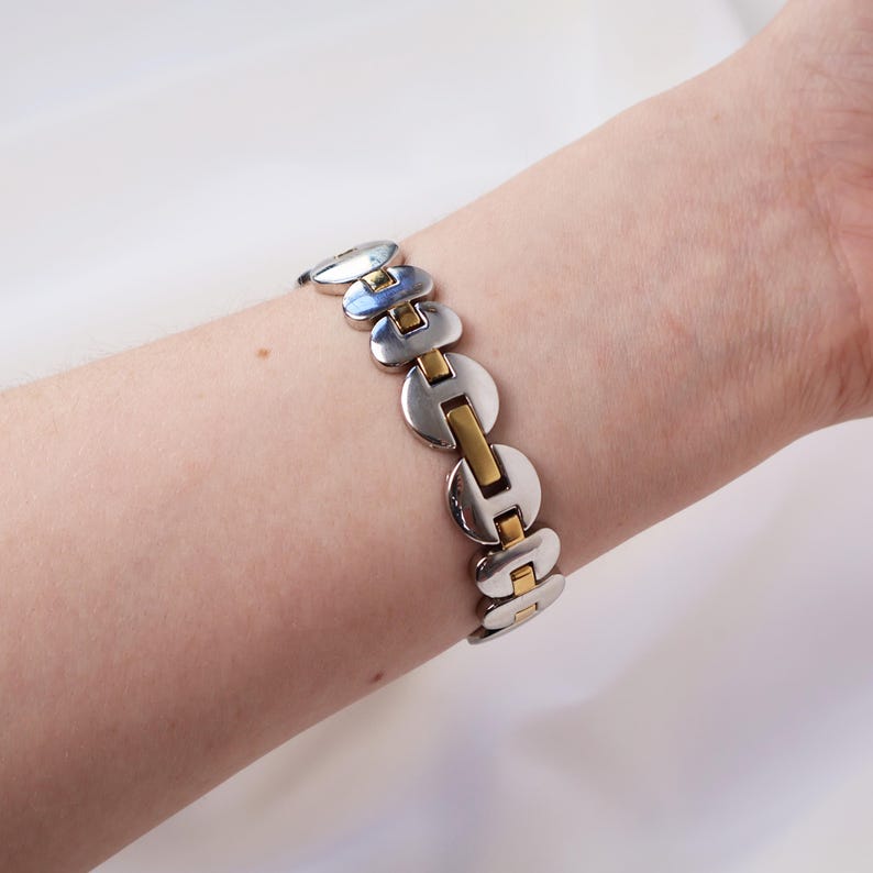 Puede incluir: Una pulsera plateada y dorada con un dise&ntilde;o de eslabones. La pulsera presenta eslabones circulares plateados conectados por barras rectangulares doradas. La pulsera se lleva en la mu&ntilde;eca.
