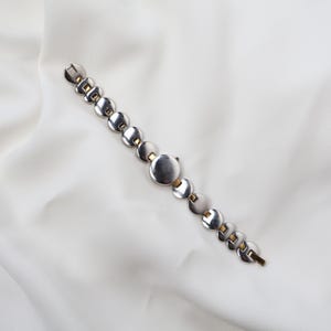 Puede incluir: Una pulsera plateada con una serie de eslabones circulares y detalles dorados. La pulsera est&aacute; dispuesta en diagonal sobre un fondo blanco. El cierre es visible al final de la pulsera.