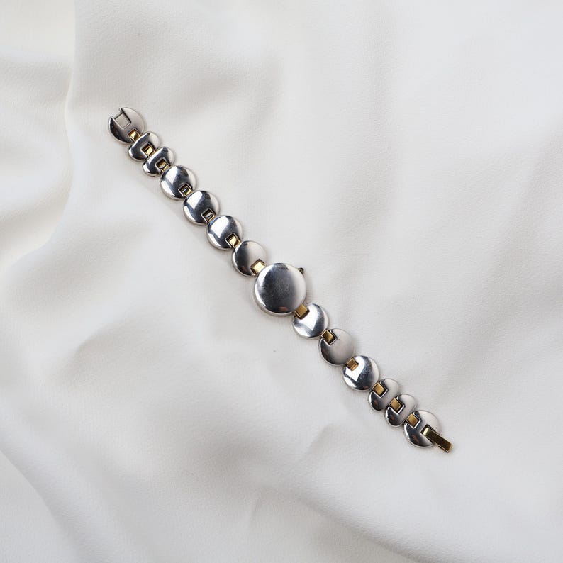 Puede incluir: Una pulsera plateada con una serie de eslabones circulares y detalles dorados. La pulsera est&aacute; dispuesta en diagonal sobre un fondo blanco. El cierre es visible al final de la pulsera.