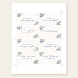 Puede incluir: Una hoja imprimible de ocho tarjetas de lugar rectangulares con fondo crema y detalles florales azules en las esquinas. Cada tarjeta muestra un nombre en letra cursiva azul, incluyendo Olivia Bennett, Grandma Rose, Sophia Martinez y Aunt Katherine.