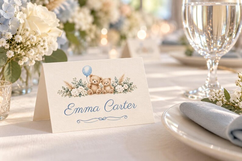 May include: A folded white place card featuring a watercolor illustration of two brown teddy bears, a light blue balloon, and white floral greenery. The name Emma Carter is printed in blue script font above a decorative blue line flourish.