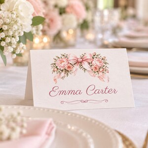 K&ouml;nnte beinhalten: Eine wei&szlig;e, gefaltete Tischkarte mit einer Aquarellillustration aus rosa Pfingstrosen und einer passenden Schleife oben. Der Name Emma Carter ist mittig in einer dunkelrosa Schreibschrift gedruckt, mit einer dekorativen Linie darunter.