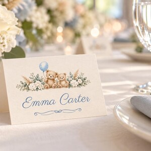 May include: A folded white place card featuring a watercolor illustration of two brown teddy bears, a light blue balloon, and white floral greenery. The name Emma Carter is printed in blue script font above a decorative blue line flourish.