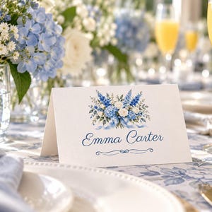 Puede incluir: Una tarjeta de sitio blanca plegada con una ilustraci&oacute;n impresa de un ramo de flores azules y blancas con una cinta azul. El nombre Emma Carter est&aacute; impreso en letra cursiva azul debajo de las flores, con un acento de l&iacute;nea azul decorativo.