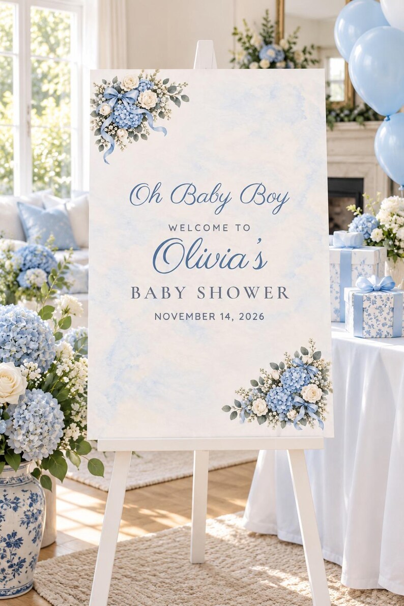 May include: A rectangular baby shower welcome sign on a white easel featuring a light blue watercolor background, floral arrangements of blue hydrangeas and white roses in corners, and text reading Oh Baby Boy Welcome to Olivia's Baby Shower November 14, 2026.