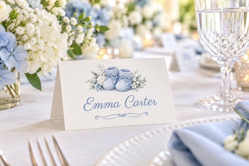 Pu&ograve; includere: Un segnaposto bianco piegato con un'illustrazione ad acquerello di scarpine da neonato azzurre circondate da piccoli fiori bianchi e blu. Il nome Emma Carter &egrave; stampato in un carattere corsivo blu sotto le scarpine con una linea.