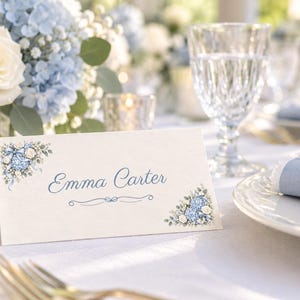 Puede incluir: Una tarjeta de sitio blanca plegada con ilustraciones florales azules en las esquinas superior izquierda e inferior derecha. El nombre Emma Carter est&aacute; impreso en letra cursiva azul en el centro, con un adorno de l&iacute;nea azul debajo.